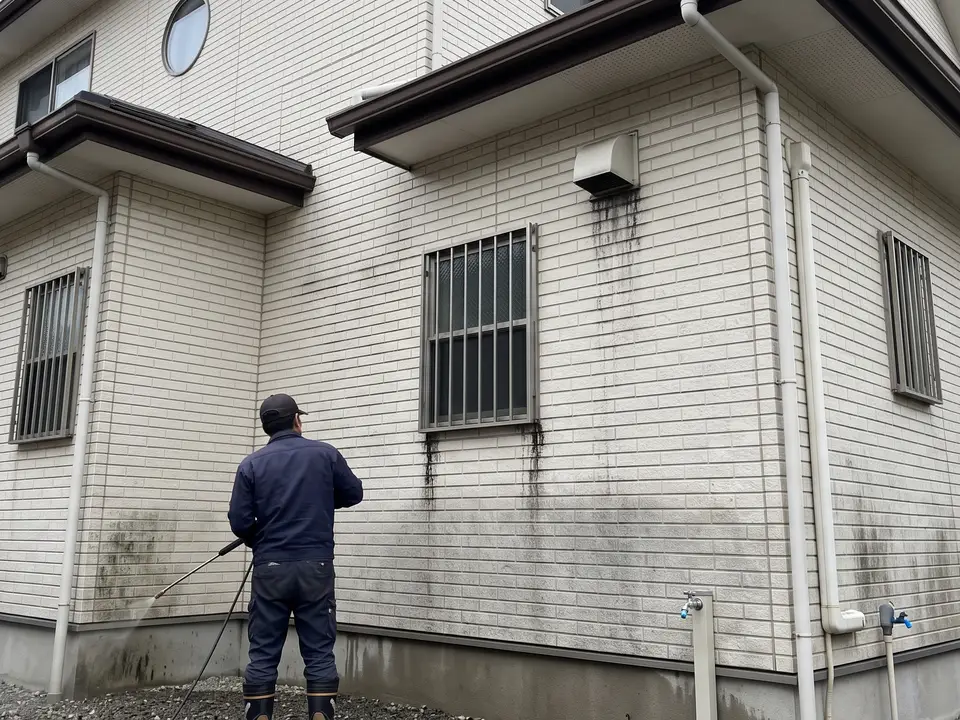 雨だれの筋と藻で黒ずんだ窯業系サイディングの外壁。職人が洗浄棒で作業している